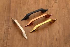 "Set of four modern metal cabinet and drawer pull handles in black, copper, gold, and silver finish placed on a wooden surface."