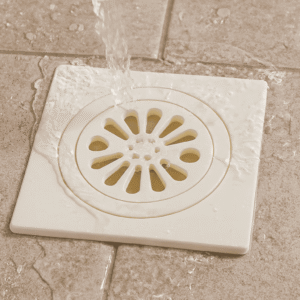 White plastic floor drain with circular decorative cover featuring radial oval and central round holes, installed on textured tiles with water flow.