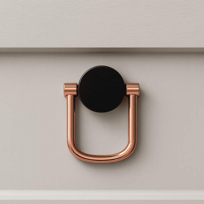 Modern U-shaped drawer pull handle with a round black base plate in rose-gold finish, mounted on a light-colored cabinet drawer.